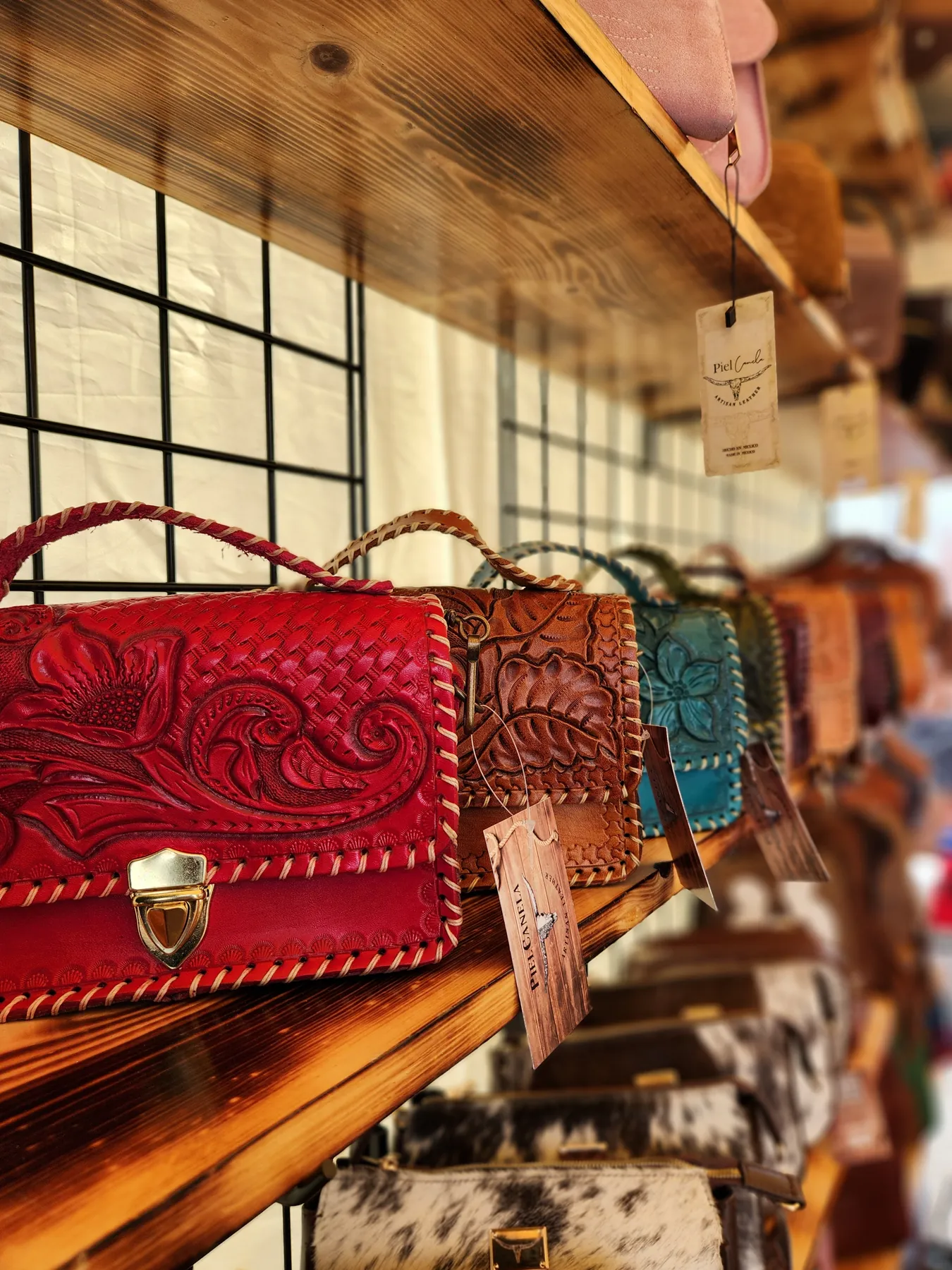 Hand-tooled leather purses in red, tan and teal on a wooden shelf, with Piel Canela tags